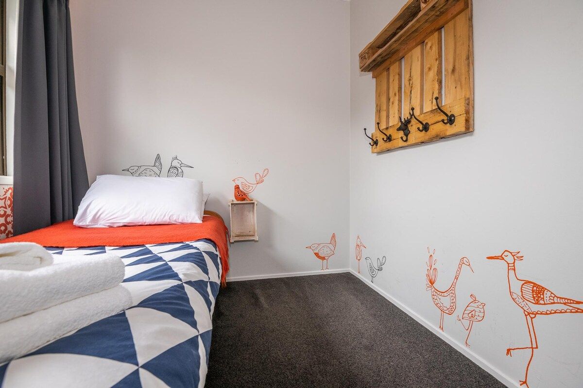 A hotel room with a bed, a small bedside table, a wall-mounted shelf, and a window with curtains.