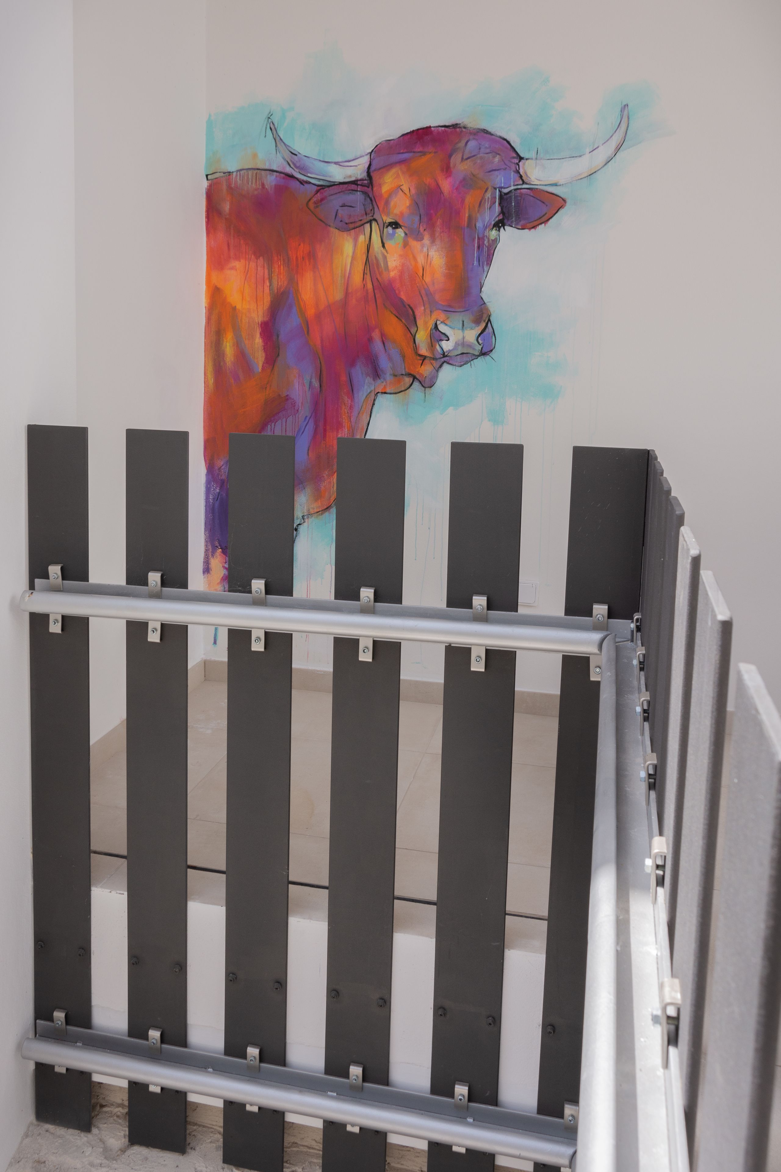 Een metalen hekwerk met verticale spijlen en een metalen leuning, dat leidt naar een ruimte met een kleurrijke muurschildering van een stier.