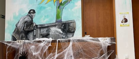 A room with a mural of a sunflower and a figure, a wooden table with a spider web, and a door in the background.