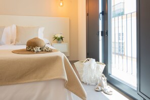 Una cama bien hecha con una manta color beige, un sombrero tejido y un bolso blanco en el suelo, al lado de una ventana con vistas a un edificio.