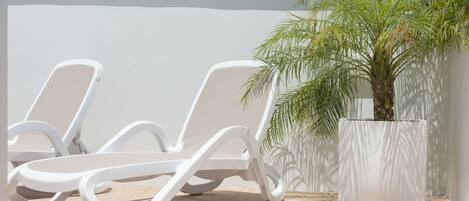 Un espacio exterior iluminado por el sol con sillones blancos y una palmera en maceta.
