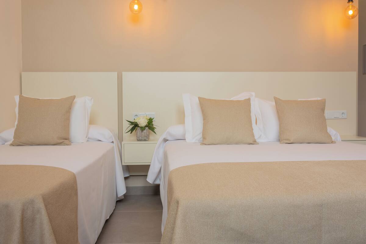A hotel room with two beds, each with a beige bedspread and pillows, a nightstand with a plant, and a wall-mounted light fixture.