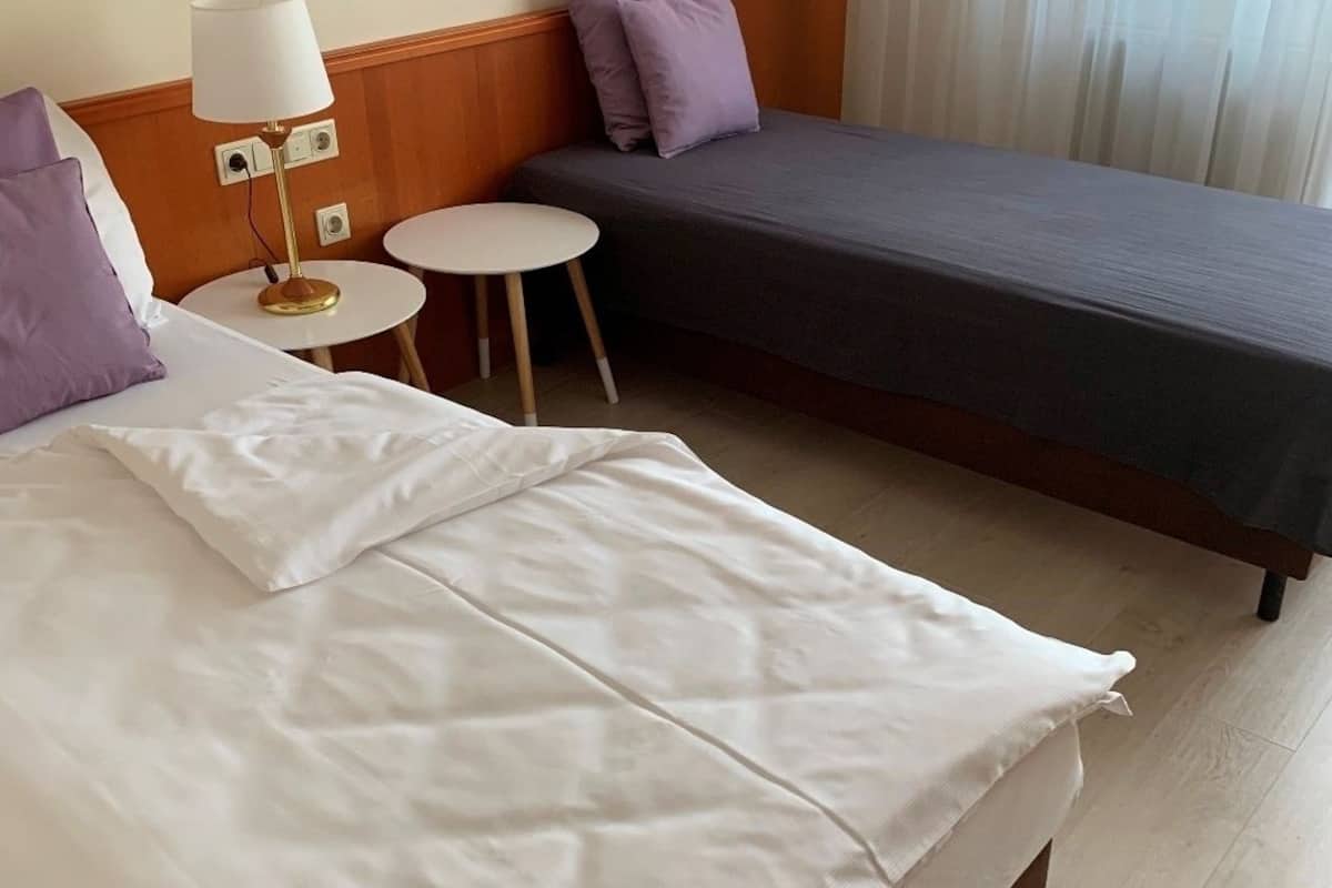 Ein Hotelzimmer mit einem großen Bett, einem Einzelbett, einem runden Tisch, einer Lampe und einem Fenster mit Vorhängen.