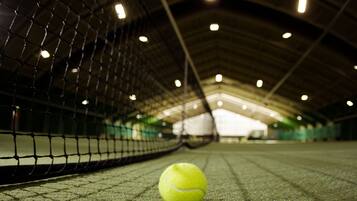 A tennis ball on a court with a net in the foreground and a roofed structure in the background.