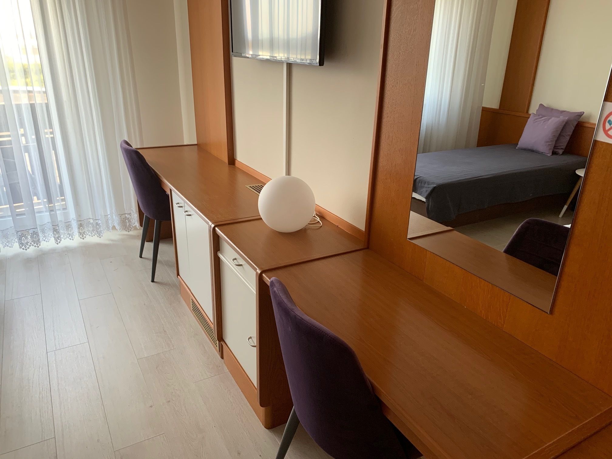 A hotel room with a wooden desk, a purple chair, a bed with a grey bedspread, and a large mirror reflecting the bed and a window with sheer curtains.