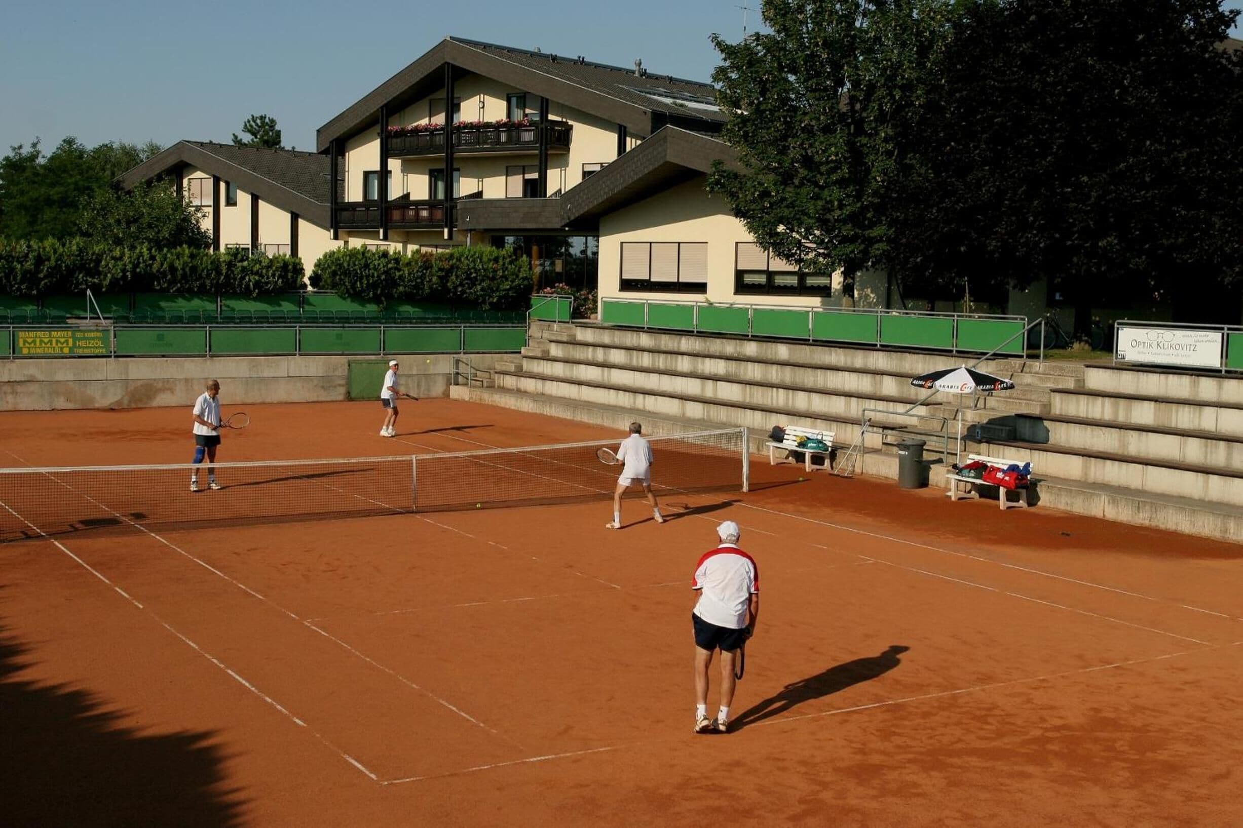 A tennis court with a net, surrounded by a building with balconies and a green fence.