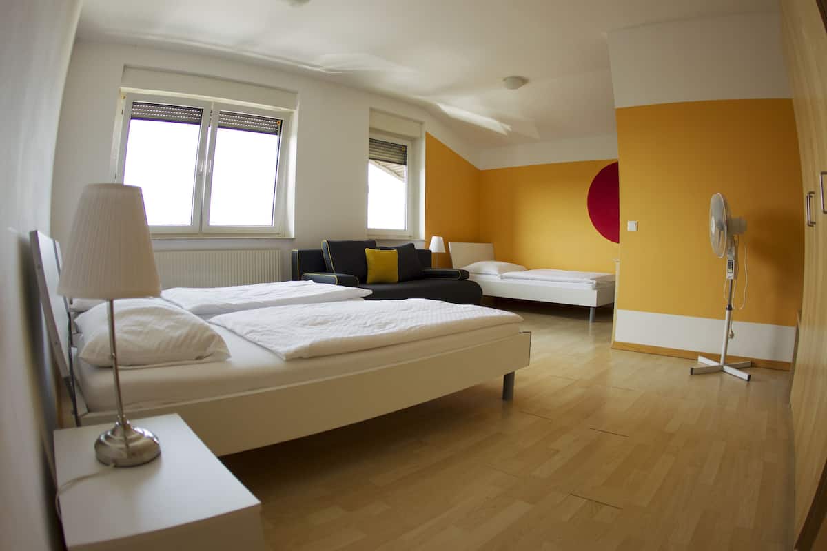Ein Hotelzimmer mit zwei Betten, einem Sofa, einer Lampe, einem Ventilator und einem Fenster mit Jalousien.