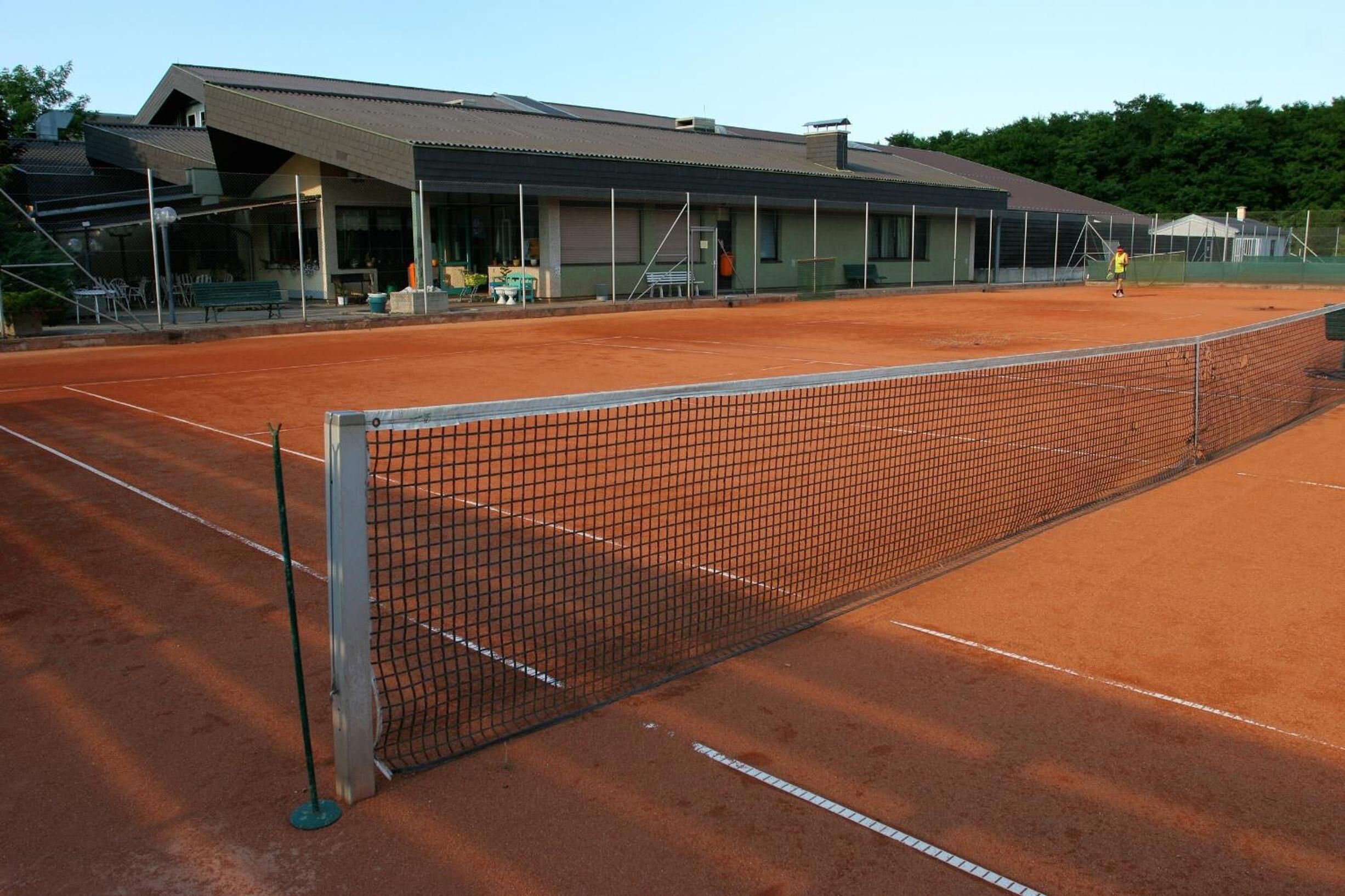 Een tennisbaan met een net, een gebouw met schuin dak en buitenzitplaatsen.