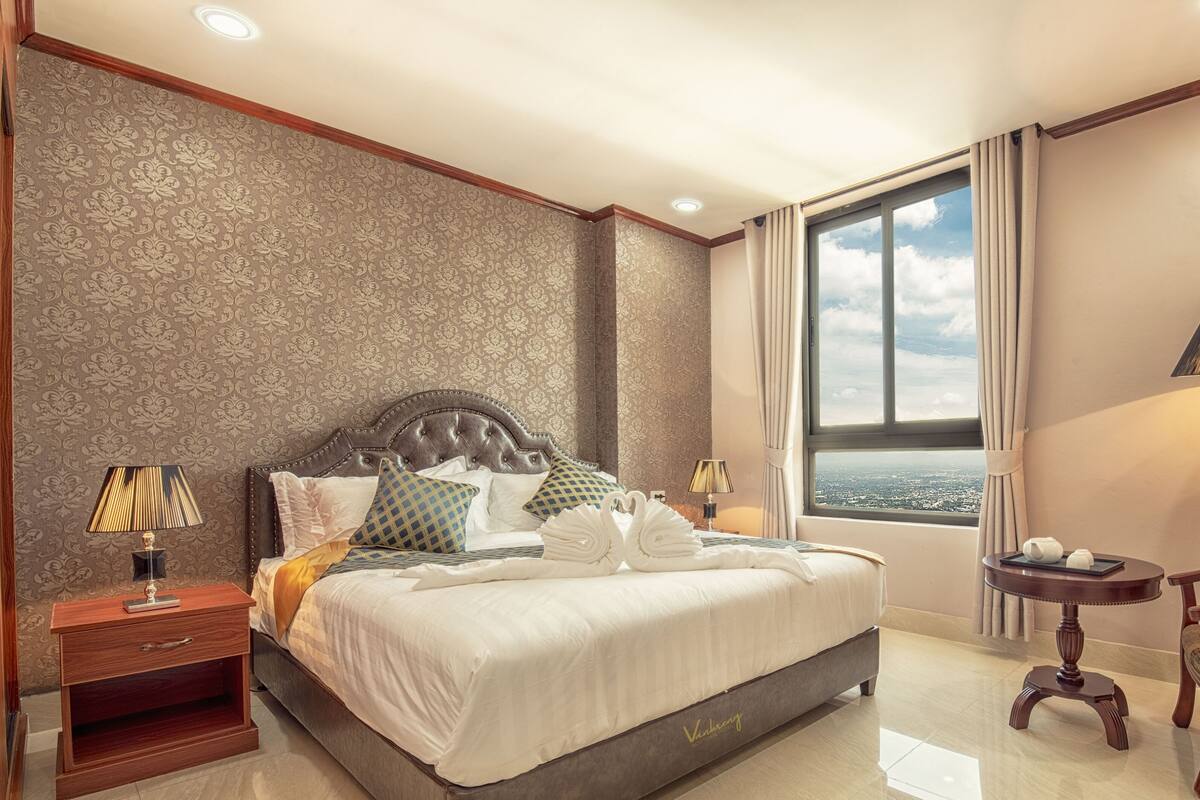 Una habitación de hotel con una cama grande, dos mesitas de noche, una silla, un ventanal con vista a la ciudad y una lámpara.