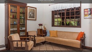 Una sala de estar con sofá, sillas, un aparador de madera con puertas de cristal, un cuadro enmarco y una ventana con cortina de encaje.