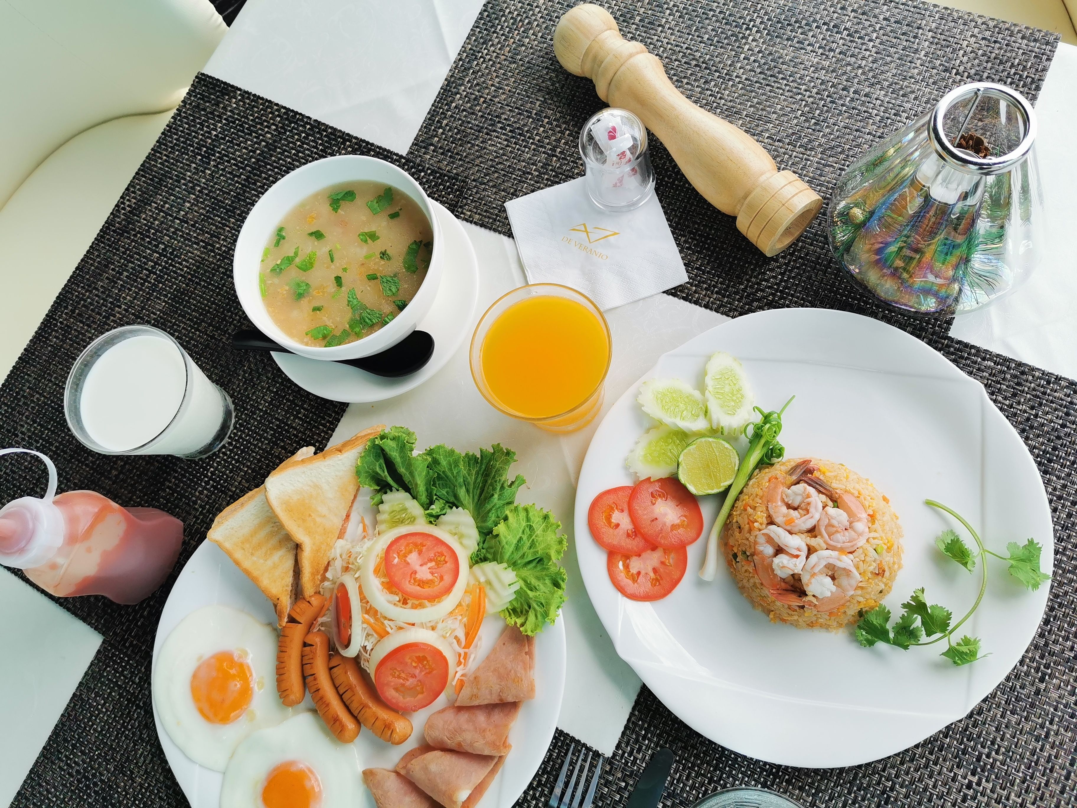 Una mesa de desayuno con un tazón de sopa, un plato de huevos, salchichas, tostadas y un plato de arroz frito con camarones.