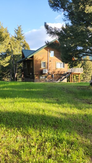 Una cabana de fusta de dos pisos amb un balcó, envoltada per un jardí amb gespa i arbres.