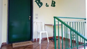 Una puerta verde con un letrero de 'B&B' arriba, una silla blanca y un felpudo con la palabra 'BIENVENIDOS' en la parte delantera.