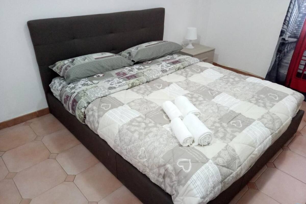 A neatly made bed with a patterned bedspread and pillows, a bedside table with a lamp, and hexagonal floor tiles.