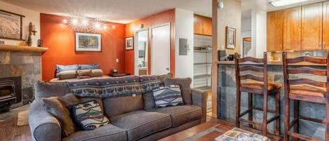 Una sala de estar con chimenea, un sofá con cojines decorativos, una mesa de centro con controles remotos y una zona de cocina visible a través de una puerta abierta.