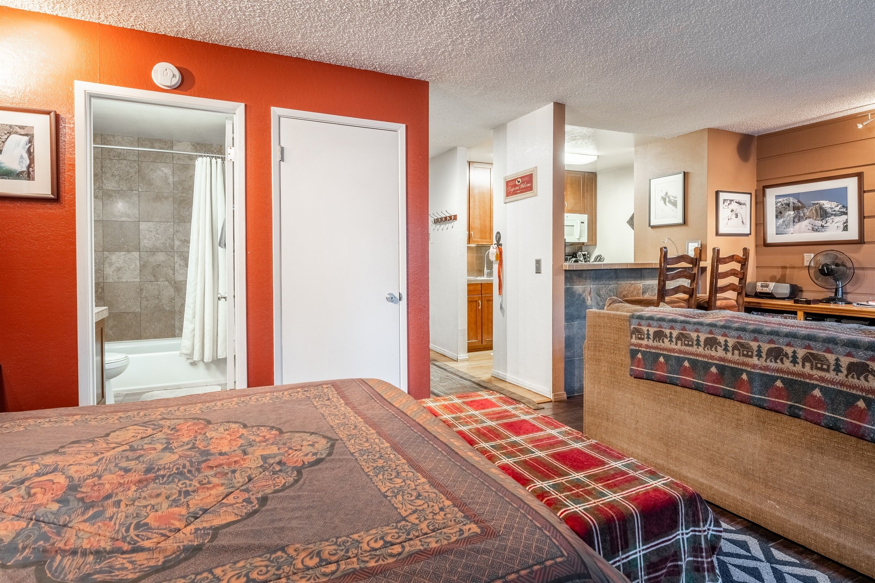 A room with a bed, a bench, a shower, a kitchen area, and a dining table.