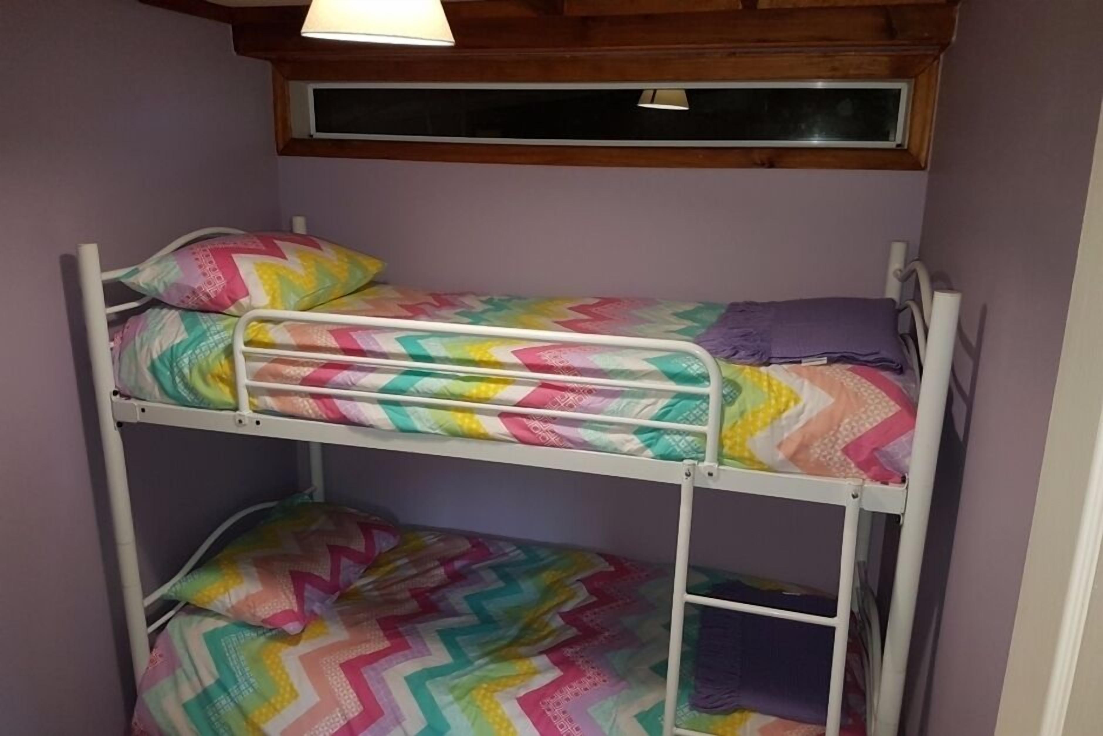A bunk bed with colorful bedding in a room with wooden ceiling beams and a window above the top bunk.