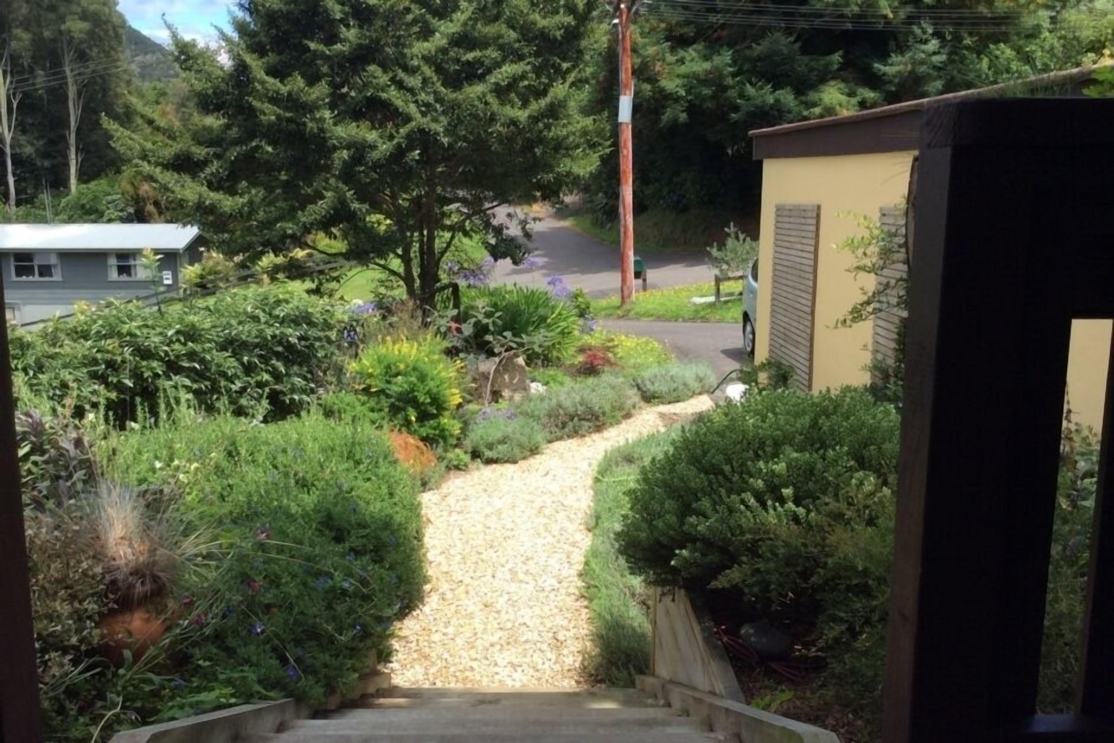 A gravel pathway leading to a house surrounded by dense greenery and a mountain in the background.