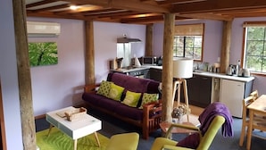 A cozy living room with a purple sofa, a small table, and a green armchair. A kitchen area is visible in the background with a microwave and a kettle. - Charming Holiday Hideaway near the Lake (Lake Okareka)
