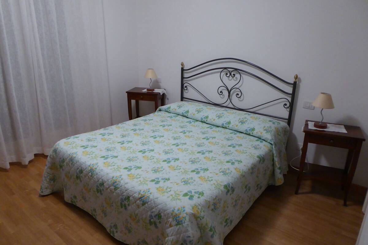 Un dormitorio con una cama, dos mesitas de noche, una lámpara sobre cada una y una cortina en la ventana.