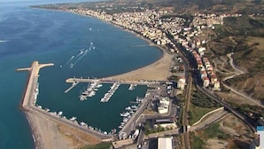 Vista aérea de un pueblo costero con un puerto deportivo, edificios residenciales y una carretera que serpentea a lo largo de la costa.