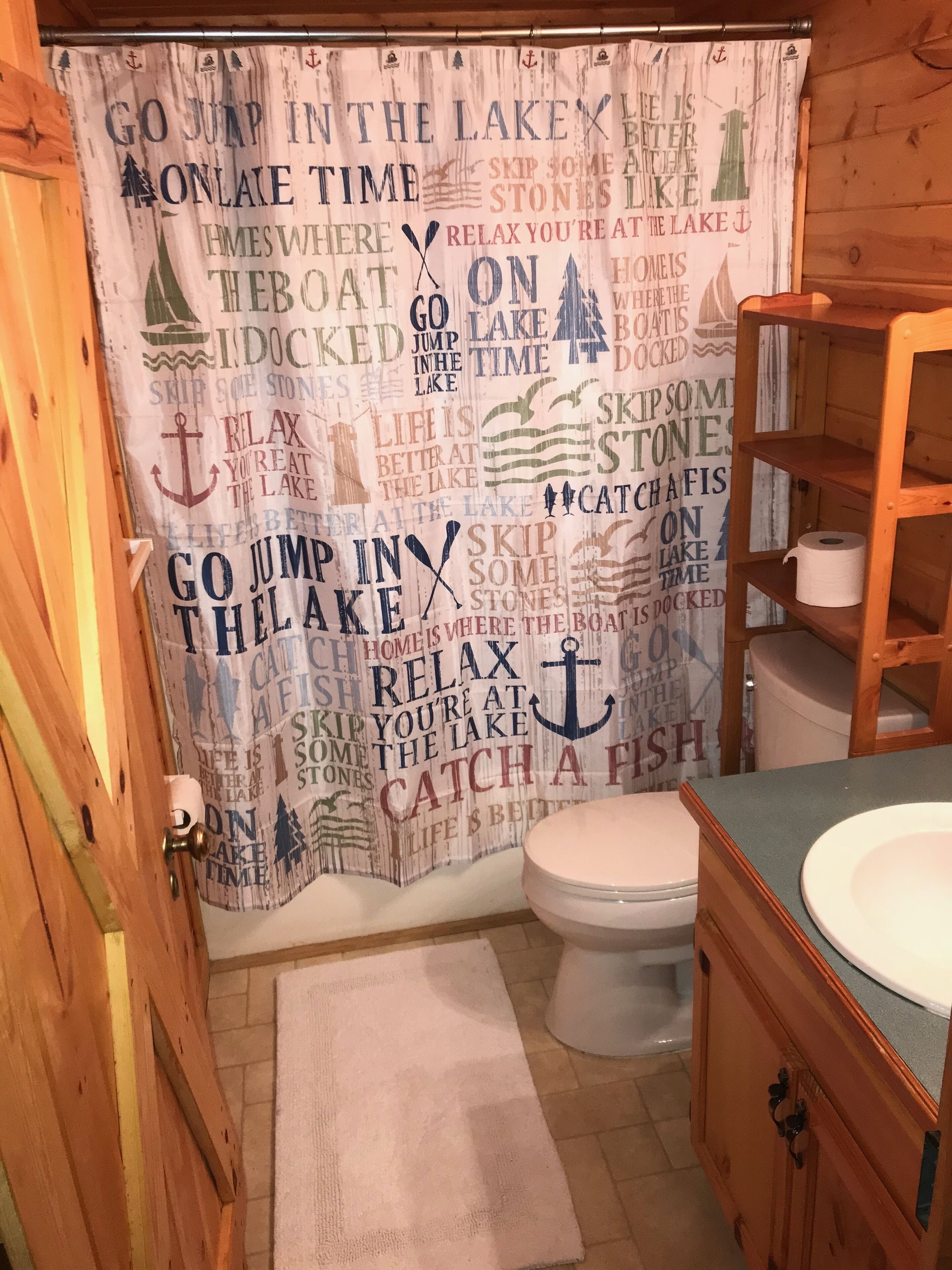 Un baño con puerta de madera, inodoro blanco, estante de madera con un rollo de papel higiénico y cortina de ducha con motivos náuticos.
