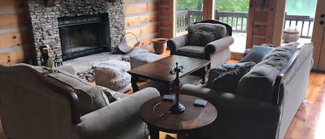 A cozy cabin interior with a stone fireplace, wooden walls, a sofa, armchairs, and a view of a lake through large windows.