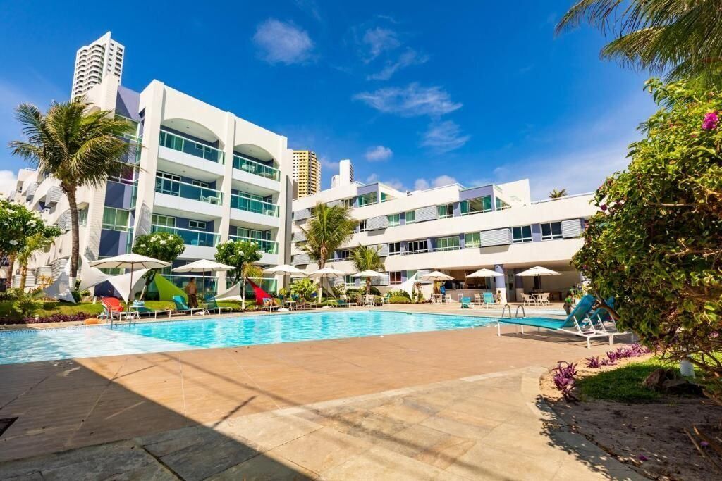 A modern hotel with a swimming pool, palm trees, and a clear blue sky.