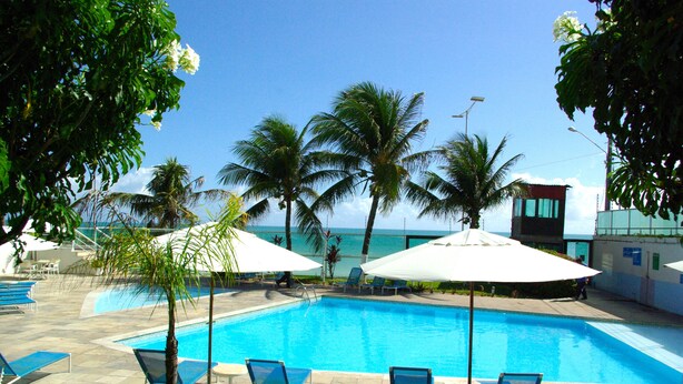 Une piscine entourée de chaises longues et de parasols, bordée de palmiers et surplombée d’un ciel dégagé.