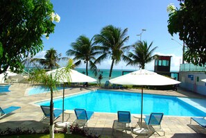 Ein Swimmingpool mit Liegestühlen und Sonnenschirmen, umgeben von Palmen und einem klaren Himmel.