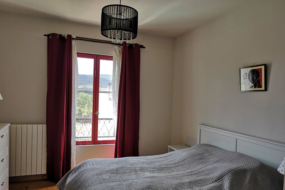 Een slaapkamer met een bed, een dressoir, een radiator, een raam met gordijnen en een kroonluchter.