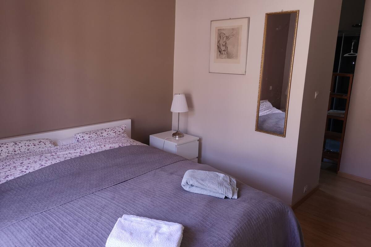 Een opgemaakt bed met een grijze plaid en witte kussens, een bedlampje, een ingelijst foto aan de muur en een spiegel.