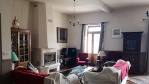 Een woonkamer met een open haard, een televisie, twee witte banken, een rode fauteuil en een boekenkast.