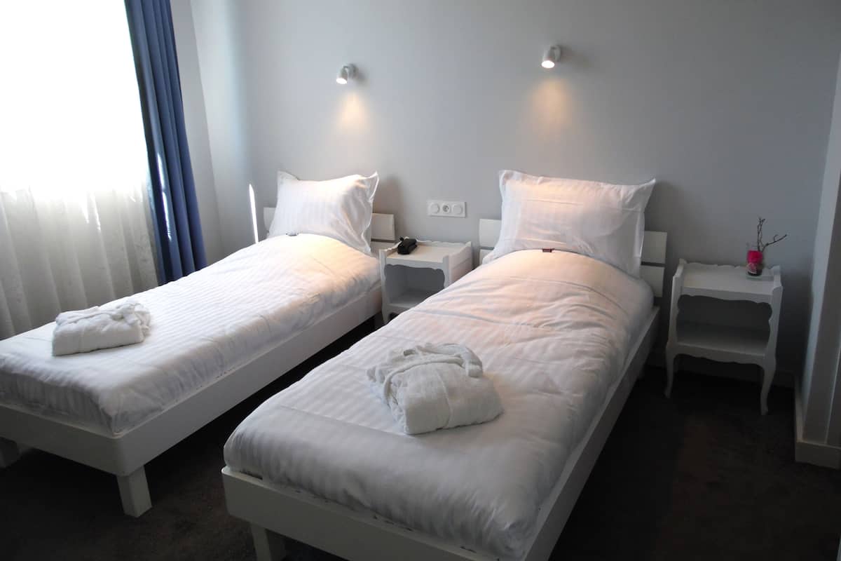 A hotel room with two single beds, each with a white duvet and pillows, bedside tables with lamps, and a window with curtains.