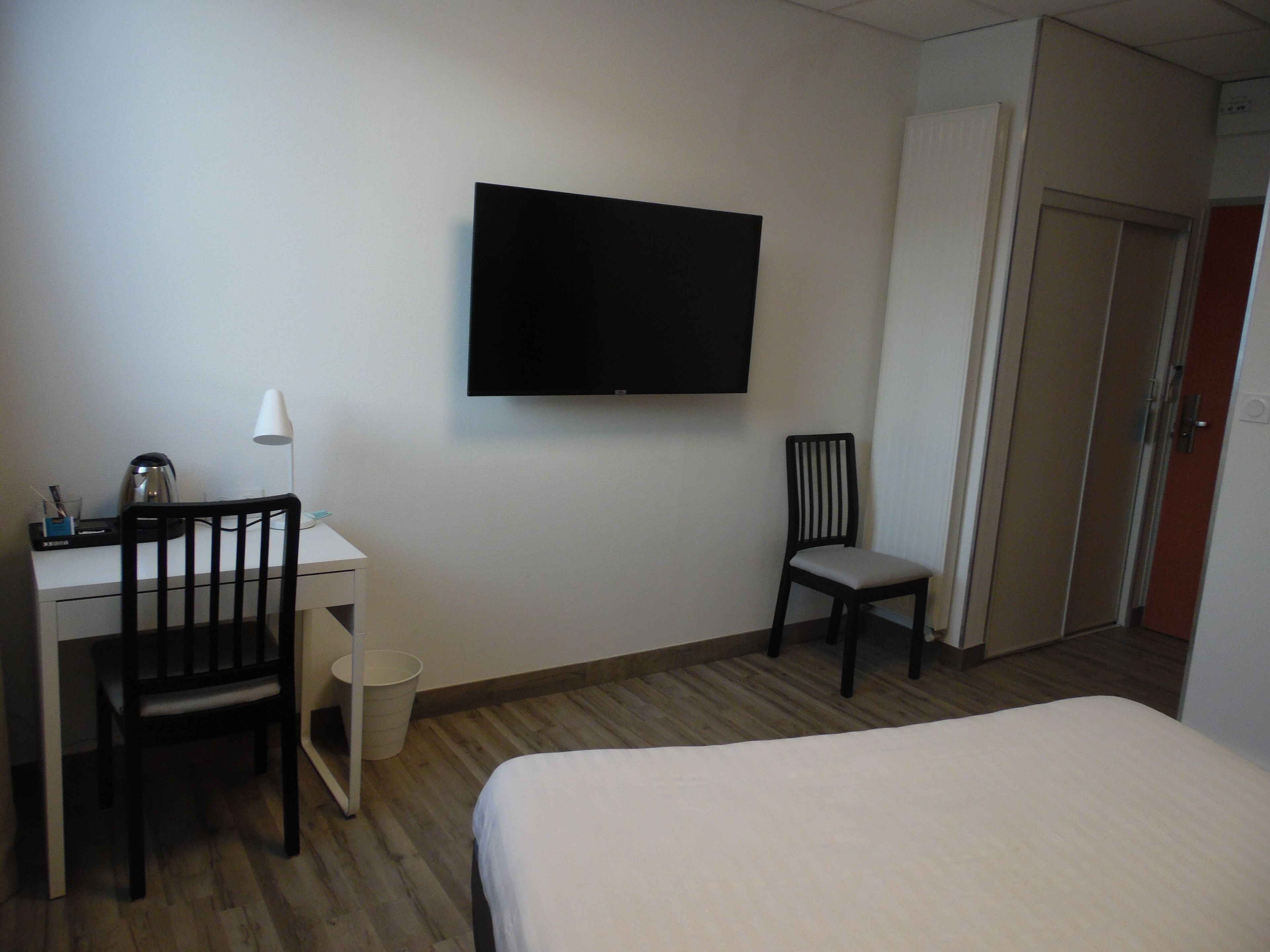 A hotel room with a bed, a desk with a chair, a television mounted on the wall, and a door leading to another room.