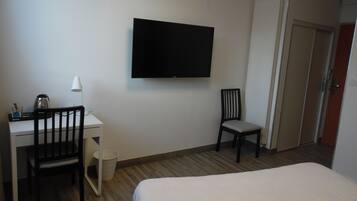 A hotel room with a bed, a desk with a chair, a television mounted on the wall, and a door leading to another room.