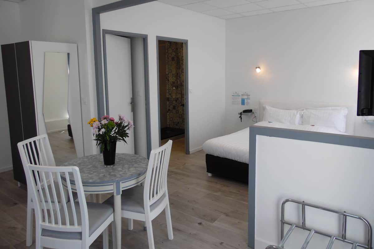 A modern hotel room with a bed, bedside table, and a small desk with a chair. There is a dining area with a table and chairs, and a bathroom visible through an open door.