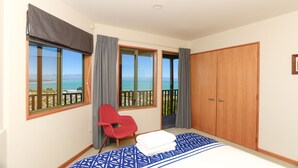 A bedroom with a bed, a red chair, a wooden wardrobe, and large windows with a view of the sea.