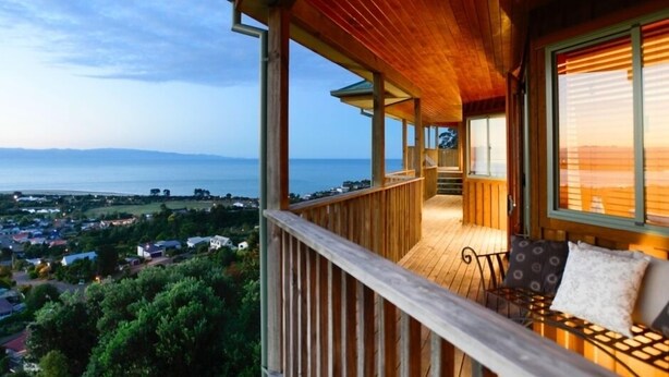 A wooden deck with a bench and a table, overlooking a coastal town and the sea.