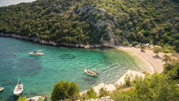 Blick auf die Küste mit Booten in klarem türkisblauem Wasser, einem Sandstrand und einer bewaldeten Hügelsicht.