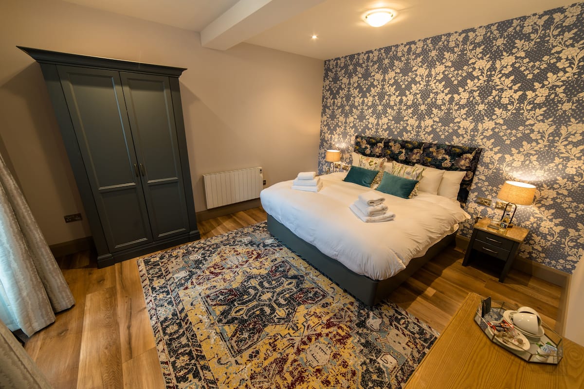 A bedroom with a large bed, dark wardrobe, floral wallpaper, and a patterned rug.
