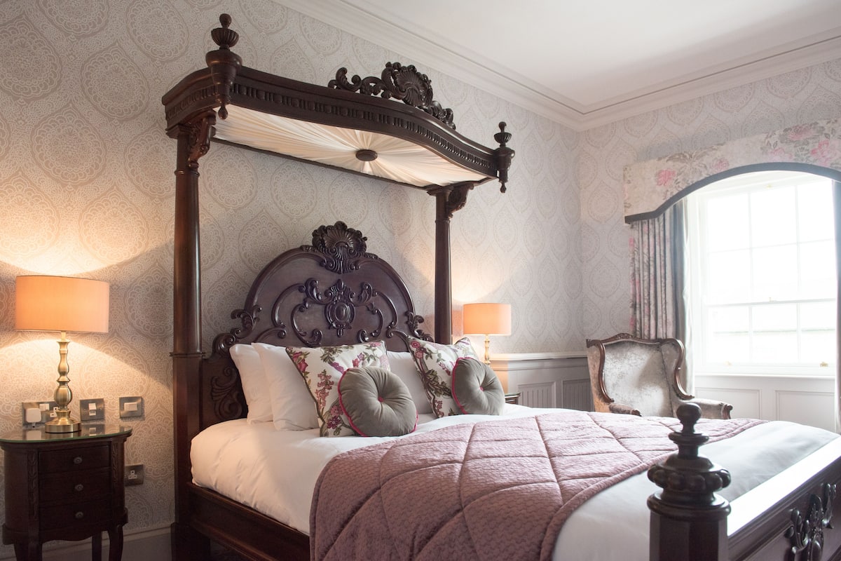 A four-poster bed with a canopy, a nightstand with a lamp, and a chair by the window.