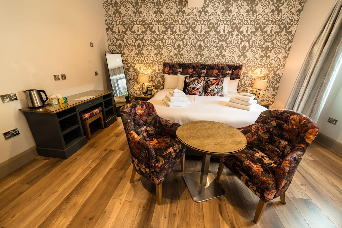 A hotel room with a large bed, two floral armchairs, a small round table, a nightstand with a lamp, and a wooden side table with a kettle and a box of tissues.