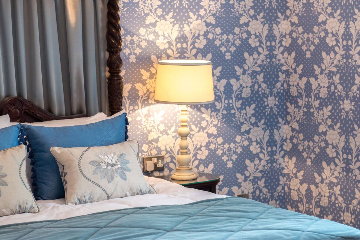 A neatly made bed with a blue quilt, a wooden headboard, and floral-patterned wallpaper.
