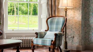 A traditional armchair with a plaid pattern, positioned next to a table and a floor lamp, in a room with floral wallpaper and a view of greenery through a large window.