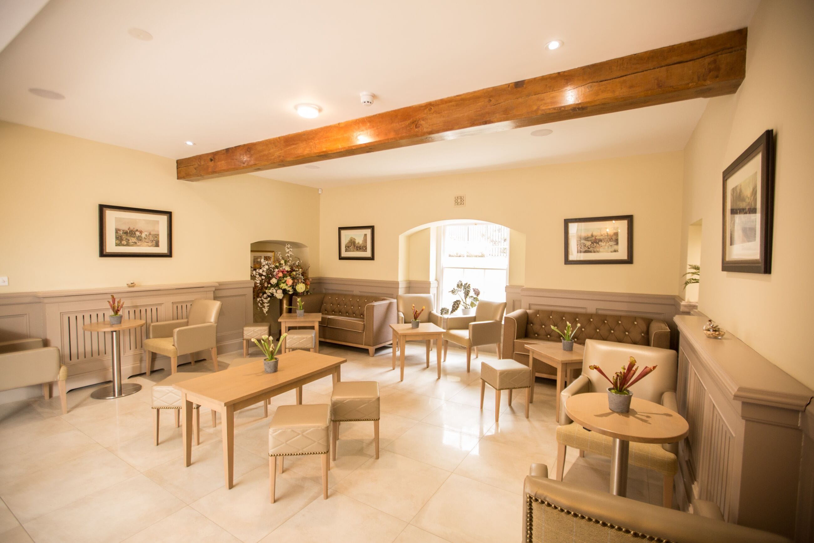 A spacious living room with a high ceiling, wooden beams, and a large window. There are multiple seating areas including sofas and chairs, and a dining table.