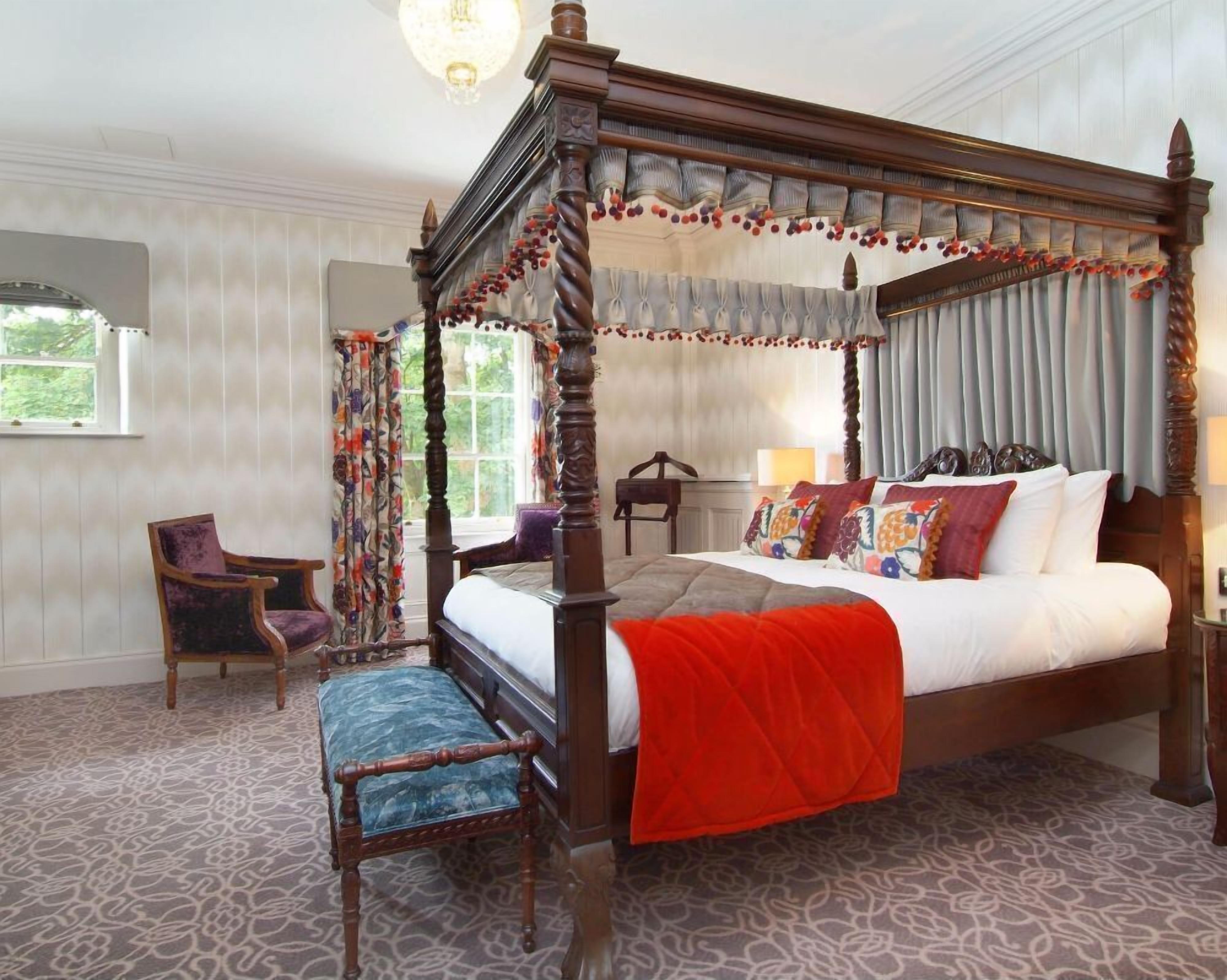 Una cama con dosel y cuatro columnas, con una manta roja, una silla de madera y una alfombra estampada.