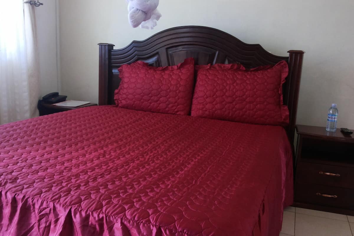 A neatly made bed with a red quilt and pillows, a bedside table with a bottle of water and a remote control, and a nightstand with a phone.