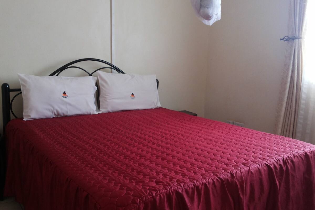 A bed with a red quilt, two white pillows with logos, a black metal headboard, and a white wall with a power outlet.