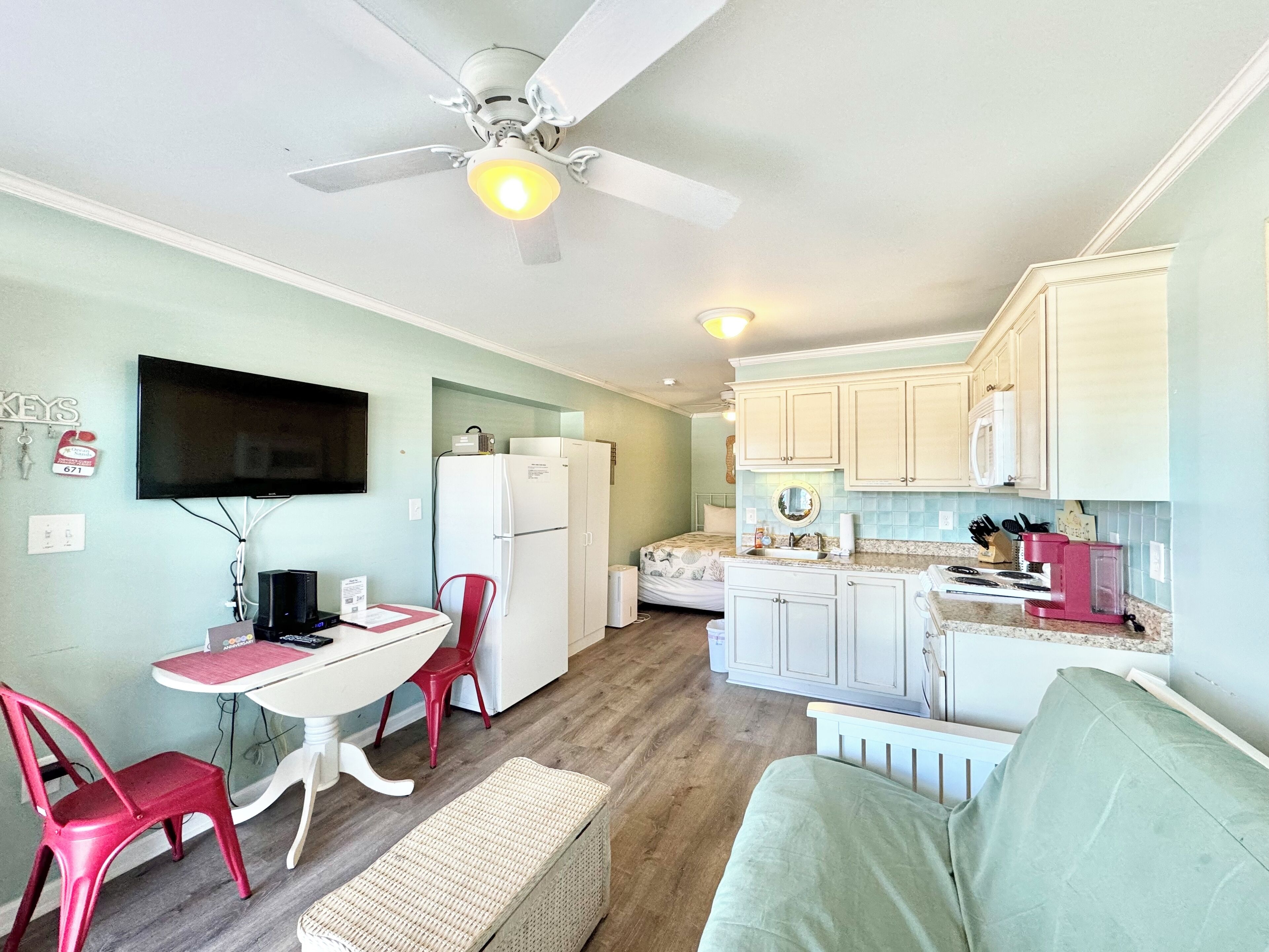 Un espace de vie compact comprenant un coin cuisine, un coin repas avec une table et des chaises, un canapé et un ventilateur de plafond.